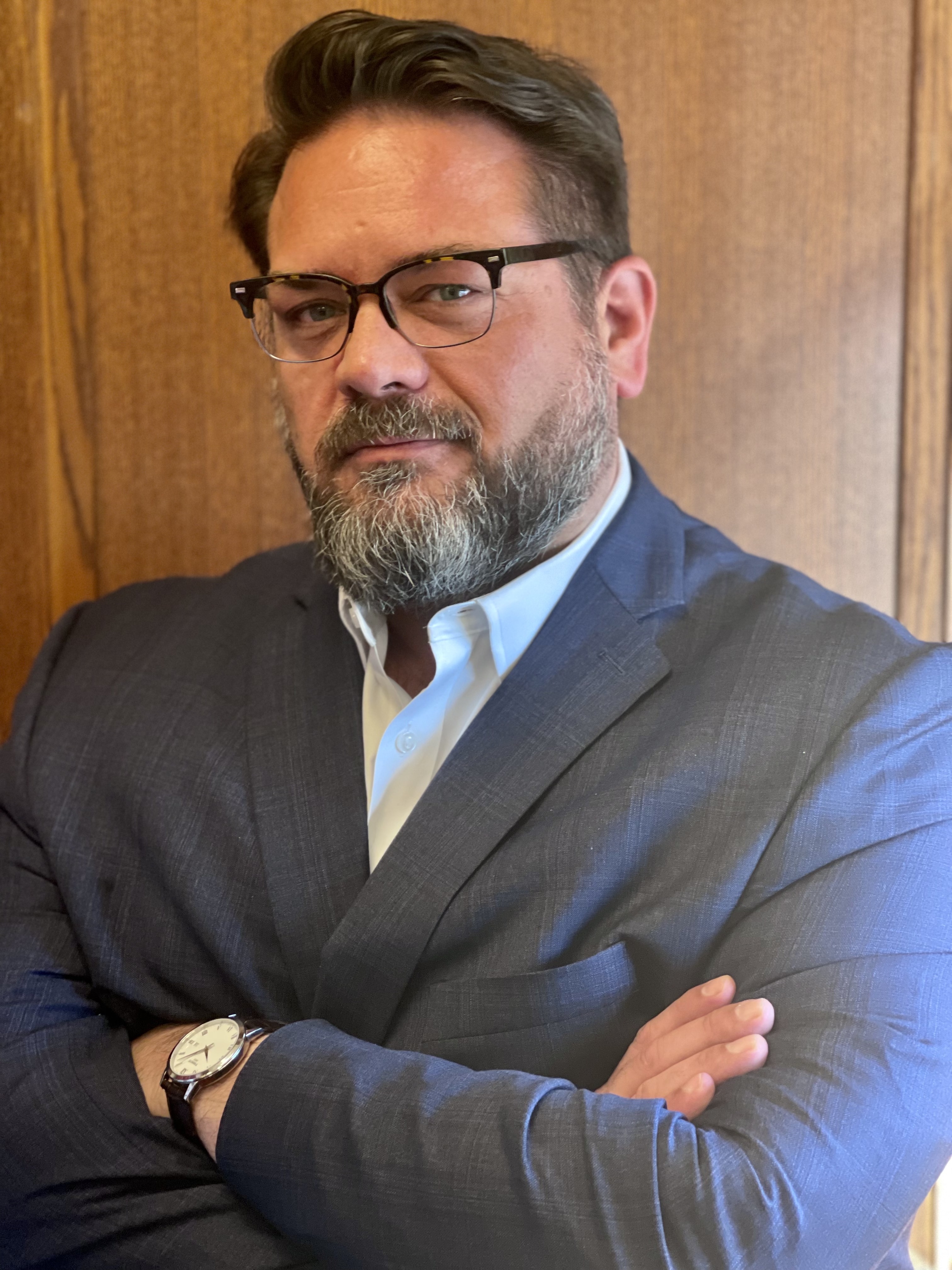 David McLeod has brown-gray hair, a beard, and glasses, and is smiling. He is wearing a light-colored collared shirt, a dark suit jacket, and a watch. He stands confidently with arms crossed against a wood-paneled background.